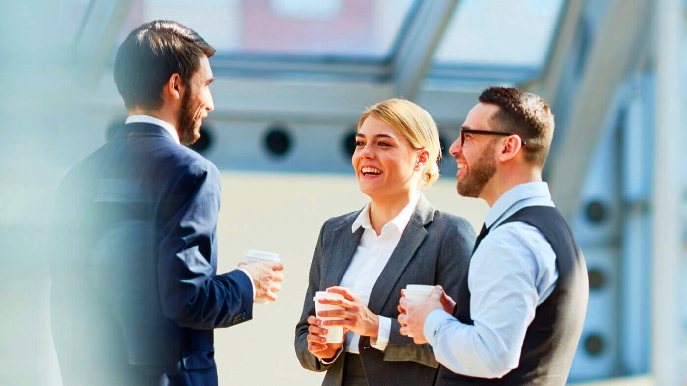 A professional setting where three business professionals, two men and one woman, are engaged in a conversation while holding coffee cups, smiling, and dressed in formal attire inside a modern office with large windows.